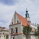 Szary budynek z czerwoną dachówką i zieloną wieżą. W dole zdjęcia grupa dzieci.