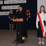 Na głównym planie przemawia mężczyzna w sutannie, stojąc przy mównicy z prawej strony dziewczyna przepasana biało - czerwoną flagą, w tle dwie kobiety stojące przy dekoracji.