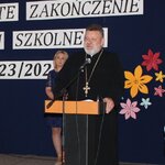 Na głównym planie przemawia mężczyzna w sutannie, stojąc przy mównicy, w tle kobieta stojąca przy dekoracji.