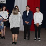 Na głównym planie znajdują się  chłopcy i dziewczęta, przed nimi dwie kobiety zwrócone przodem: jedna trzyma tacę, w tle flaga biało - czerwona.