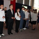 Na głównym planie stoją dziewczęta i chłopcy, trzymający w dłoniach ozdobne płaskie pudełeczka, przed nimi dwie kobiety zwrócone przodem, w tle flaga biało - czerwona i stojąca postać kobiety.