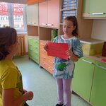 Na zdjęciu stoi dziewczynka, trzyma czerwoną kartkę z napisami dzień dobry w 3 różnych językach. Przed dziewczynką stoi chłopiec patrzy na kartkę. Tle pomarańczowo-zielone szafki.