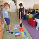 Na zdjęciu widnieje 5 dzieci. Dwóch chłopców na pierwszym planie stoi i patrzy w dół na flagi różnych krajów, rozłożone na podłodze. Jeden z chłopców w czarnej koszulce z krótkim rękawkiem wskazuje wyciągniętą ręką i palcem wskazującym na flagę Chin.  W tle siedzi trzy dziewczynki na kolorowych pufach pochylające się nad kartką.