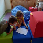 Na zdjęciu widnieje dwóch chłopców siedzących i pochylających się nad kartką. Chłopcy znajdują się w sali wśród jasno-zielonych, czerwonych i niebieskich kwadratowych puf. Chłopcy klęczą przy jednej z niebieskich puf, rozwiązują zadania na kartce.