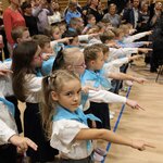 Dzieci ubrane na galowo w błękitnych chustach wyciągają prawą rękę z dwoma palcami w kieru.jpg