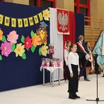 Dziewczynka stoi przed statywem_ za nią zawieszona jest dekoracja i stoi poczet sztandarow.jpg