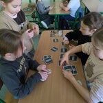 Na zdjęciu widać pięć dziewczynek i jednego chłopca siedzących przy dwóch ławkach. Jedna dziewczynka stoi przy ławce. Wszystkie osoby grają w karty na ławkach. W tle widać fragment ściany i drzwi z kolorowym plakatem.