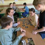 Na pierwszym planie widać dwie dziewczynki i jednego chłopca w szarej bluzie siedzących przy ławce, jeden chłopiec w czarnym T-shircie stoi przy tej ławce. Na ławce leży opakowanie od gry. Dzieci grają w memory rozłożonym na ławce. W tle widać jedną dziewczynkę i dwóch chłopców siedzących przy ławce, na której leży gra. W tle widać dwie tablice korkowe, na których zawieszone są prace plastyczne.