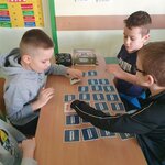 Na pierwszym planie widać czterech chłopców. Jeden z nich na nas patrzy i się uśmiecha. Drugi w szarej bluzie z kapturem oraz chłopiec w okularach grają w memory. Jeden chłopiec obserwuje grę. Wszyscy siedzą przy ławce, na której leżą też: opakowanie od gry i piórnik. W tle widać fragment ściany i drzwi z plakatem.