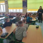 Na zdjęciu widać siedemnaścioro dzieci, którzy siedzą w parach przy ławkach. Na ławkach leżą piórniki. Po prawej stronie stoi kobieta, która zwrócona jest w kierunku tablicy interaktywnej. Widać także biurko, na którym leżą gry planszowe, a także szafki z pomocami dydaktycznymi. Mężczyzna z aparatem siedzi po prawej stronie przy ławce i przygląda się stojącej kobiecie. W oddali widać trzy okna, z których dwa są częściowo przysłonięte roletami.