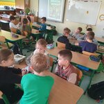 Na zdjęciu widać czternaścioro dzieci, którzy siedzą przy ławkach w czterech grupach. Wszyscy grają w karty. Na pozostałych dwóch ławkach nie ma nikogo, ale leżą cztery piórniki. Na końcu widać ścianę, na której wisi tablica interaktywna, na której prezentowana jest prezentacja oraz tablica suchościeralna z żółtą gąbką. Widać też szafkę.