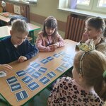 Na pierwszym planie widać uśmiechniętego chłopca w granatowej bluzie, który wraz z trzema dziewczynami gra w memory na ławce. W tle widać dwie ławki, na jednej widać fragment plecaka, a na drugiej książki z lampką. Widać także dwa parapety i fragment okna i ściany.