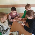 Na zdjęciu widać dwóch chłopców: jeden w brązowej bluzie, drugi w zielonej oraz dwie dziewczynki: jedna w białym futerku, druga w czarnej bluzie. Dwoje dzieci gra w dobble, jeden chłopiec w zielonej bluzie próbuje wziąć jedną kartę. Dziewczynka w białym futerku obserwuje grę pozostałych osób. Na ławce, przy której siedzą dzieci widać fragment pudełka od gry. W tle widać fragment ściany sali oraz piórnik leżący na innej ławce.