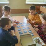 Na pierwszym planie widać dwie dziewczynki i dwóch chłopców, którzy grają w memory. Chłopiec rozdaje karty, dziewczynka trzyma kartę. Na ławce widać leżące opakowanie od gry. Widać dwa parapety i fragment ściany na drugim planie.