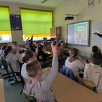 Na zdjęciu widać osiemnaścioro uczniów siedzących przy ławkach w parach. Piątka z nich ma rękę podniesioną w górze. Patrzą w kierunku wyświetlonej prezentacji na tablicy interaktywnej. Przed ławkami stoi kobieta z ręką wskazującą na uczniów. Na dalszym planie widać okna z roletami, szafki.