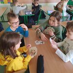 Na pierwszym planie widać dwie dziewczynki i dwóch chłopców, którzy grają w karty przy ławce. Na ławce leży też czarny piórnik. Na drugim planie widać ośmioro dzieci, którzy grają w memory przy dwóch ławkach. Dodatkowo po lewej stronie widać ławkę, na której leży zielono-czarny piórnik, a pod nią stoi niebieski we wzorki plecak oraz zauważalny jest też fragment drugiej ławki, po prawej także widać fragment jednej ławki.