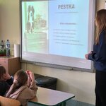 Na zdjęciu widać stojącą kobietę, która patrzy w kierunku tablicy interaktywnej. Na tablicy widać wyświetloną prezentację o psie o imieniu Pestka. Widać także dwie osoby, w tym jedną dziewczynkę patrzącą na ekran i siedzących przy ławce.