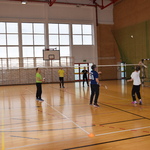 Cztery osoby grają w badmintona na hali sportowej. Obok boiska stoi sędzia. Grający chłopiec w białej koszulce przymierza się do odbicia lotki. 