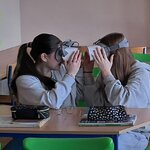 W sali lekcyjnej dwie uczennice w goglach VR siedzą w ławce zwrócone przodem do siebie. Na ławce leżą książki i piórniki.  