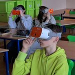 Troje uczniów siedzi w sali lekcyjnej. Na głowach założone mają gogle VR. Patrzą przez nie w różne strony.