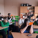 Grupa uczniów siedzi w sali lekcyjnej. Na głowach założone mają gogle VR. Patrzą przez nie w różne strony.