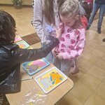 Dziewczynka w różowej bluzie z serduszkami stoi przy stoliku i przyjmuje papierową figurkę od osoby w czarnej kurtce. Na stole znajdują się tace z kolorowymi wycinankami zwierząt. Obok stoi starsza dziewczyna w jasnej bluzie z kapturem, a w tle widać innych uczestników wydarzenia w szkolnym korytarzu.