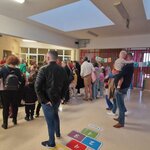 Szkolny korytarz wypełniony ludźmi – rodzice, dzieci i nauczyciele zgromadzeni na wydarzeniu. W tle widoczne są dekoracje, w tym kolorowy napis na girlandzie. Jeden z mężczyzn trzyma na rękach małe dziecko, a inni stoją lub rozmawiają. Na podłodze widać klasy do gry w klasy.
