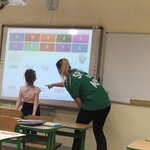 Na zdjęciu widać salę lekcyjną, w której nauczycielka i uczennica pracują przy tablicy interaktywnej. Tablica wyświetla ćwiczenie związane z liczbami i ich nazwami w jeżyku angielskim. Dziewczynka w jasnej bluzce i ciemnych spodniach stoi przy tablicy, podczas gdy nauczycielka w zielonej koszulce pochyla się i wskazuje na ekran pomagając w zadaniu.