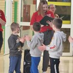 W szkolnym korytarzu nauczycielka w czerwonej koszulce trzyma czarną teczkę i uśmiecha się, czytając na głos. Przed nią stoi kilku uczniów w szarych bluzach i dresach, którzy klaszczą w dłonie. W tle widać inną nauczycielkę oraz szkolne drzwi i ściany w kolorach zieleni i żółci.