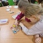Na zdjęciu dwie dziewczynki pochylają się nad małymi pojemnikami, wybierając elementy do swojej pracy. W tle widać zielone krzesła oraz maszyny do szycia. zajęcia odbywają się w pracowni technicznej.
