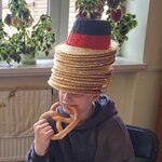 Na zdjęciu widać chłopaka, który siedzi na krześle i je precla. Na głowie ma wiele kapeluszy nałożonych jeden na drugi. Są one w kolorze czarno-czerwono-złotym.