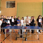 Grupa młodzieży i dorosłych stoi obok siebie w hali sportowej.  Przed nimi stoi stół do tenisa. Niektórzy trzymają w rękach dyplomy. Wszyscy ubrani są w stroje sportowe. 