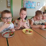 Na głównym planie widać czterech chłopców, którzy siedzą przy zsuniętych ławkach szkolnych. Chłopiec pierwszy od lewej strony gryzie kanapkę nad swoim talerzem. Drugi chłopiec od lewej trzyma w ręku kanapkę i na nią patrzy. Trzeci chłopiec od lewej jest obejmowany ręką przez ostatniego chłopca, który się uśmiecha i patrzy przed siebie. W tle widać tablicę korkową z pracami plastycznymi na niebieskim tle oraz biurko, krzesło i lampkę.
