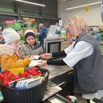 na zdjęciu widać dwójkę dzieci: dziewczynka w żółtej kurtce z puszkiem i w białej czapce oraz chłopca w szarej kurce i czapce w paski. Dziewczynka i chłopiec przyjmują paragon od pani kasjerki stojącej za kasą. Z lewej strony widać także kosz z musami i czekoladami. W tle widać mężczyznę stojącego tyłem oraz inny asortyment sklepu spożywczego.