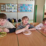 Na zdjęciu widać trzech chłopców, dziewczynkę i fragment drugiej dziewczynki. Chłopiec pierwszy od lewej strony je kanapkę nad żółtym talerzem pełnym warzyw. Drugi od lewej chłopiec uśmiecha się i patrzy przed siebie trzymając kanapkę nad talerzem z chlebem posmarowanym pasztetem. Dziewczynka z dłuższą grzywką w jasnej bluzie siedzi przy stole, na którym stoi różowy bidon.  Jeden chłopiec w granatowym T-shircie stoi w tle i ma otwartą buzię. W tle widać dwie tablic z pracami plastycznymi uczniów, fragment ściany i plecaki leżące na podłodze.