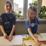 Na głównym planie widać dwie dziewczynki. Dziewczynka z lewej strony jest wyższa, kroi ser żółty na desce do krojenia i uśmiecha się. Druga dziewczynka w niebieskim T-shircie kładzie ser na talerz, też jest uśmiechnięta, ale nie patrzy do przodu. W tle widać kwiaty na parapecie, talerze z chlebem i fragment okna.