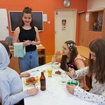 Na zdjęciu widać salę lekcyjną, a w niej trzy dziewczyny siedzące przy stole. Na stole stoją naczynia, szklanki z napojami i butelka oraz jedzenie zrobione z papieru. Obok stołu stoi dziewczyna ze ścierką kuchenną przewieszoną przez rękę. W dłoni trzyma długopis.