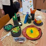 Na zdjęciu widać stół z serwetą, a na nim naczynia i dania wykonane z papieru, serwetki oraz plastikowe sztućce. Obok stołu stoi dziewczyna, która trzyma w ręce kartę dań w języku niemieckim.