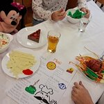  Na zdjęciu widać stół z naczyniami, papierowe potrawy, kartę dań  w języku niemieckim, dwie szklanki z napojami i fragmenty trzech osób siedzących przy stole.