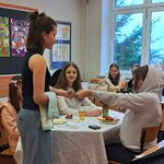 Na zdjęciu widać stół z jedzeniem wykonanym z papieru oraz szklanki z napojami. Przy stole siedzą trzy dziewczyny. Jedna z nich podaje coś dziewczynie stojącej obok. W tle widać dwie inne dziewczyny siedzące przy stole.