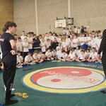 Grupa zawodników siedząca dookoła maty podczas rozpoczęcia zawodów słuchając informacji od sędziów turnieju.