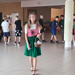 Po holu szkolnym idzie dziewczyna ubrana w sukienkę bawarską, a w ręce trzyma koszyczek. W tle widać uczniów.