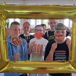 Na zdjęciu widać fotobudkę w formie złotego prostokątnego balona, a w niej sześciu chłopców trzymających symbole niemieckie – kiełbaskę, wąsy w kolorach flagi Niemiec, precle, rudą brodę, rude warkocze, kapelusz i sukienkę bawarską.