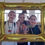 Na zdjęciu widać fotobudkę w formie złotego prostokątnego balona, a w niej trzy uśmiechnięte dziewczyny trzymające symbole niemieckie – precle, wąsy w kolorach flagi Niemiec oraz kapelusz bawarski.