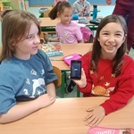 Dwie dziewcznki siedzą przy stoliku_ jedna prezentuje starszy typ telefonu.jpg