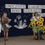 Trzy kobiety stoją na tle granatowej dekoracji z napisem „Uroczyste zakończenie roku szkolnego 2024/2025”. Jedna z nich, ubrana w biało-niebieską sukienkę, dzwoni dzwonkiem, symbolicznie kończąc rok szkolny. Obok niej duża dekoracja z żółtymi słonecznikami w czarnej donicy. W tle wy-cinanki przedstawiające dmuchawce i liście, a przed kobietami stoi mikrofon