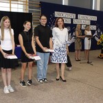 Czworo uczniów stoi na tle sali gimnastycznej podczas uroczystego zakończenia roku szkolnego 2024/2025. Troje z nich trzyma nagrody książkowe. Obok nich stoi kobieta w białej sukience z nie-bieskim wzorem. W tle dekoracja z napisem „Uroczyste zakończenie roku szkolnego 2024/2025”, motywami dmuchawców i dużymi sztucznymi słonecznikami w donicy. Dwie inne kobiety prowadzące uroczystość stoją przy mikrofonie