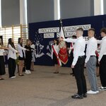 Uczniowie podczas uroczystości zakończenia roku szkolnego 2024/2025 składają przysięgę. Grupa młodzieży stoi w szeregu, z wyciągniętą ręką w geście przysięgi. W centrum stoi uczennica i ucznio-wie - poczet sztandarowy ze sztandarem Polski. W tle widoczna dekoracja z napisem „Uroczyste za-kończenie roku szkolnego 2024/2025”, polska flaga i kwiaty słoneczników.