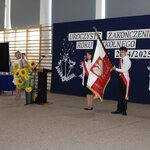 Uroczyste zakończenie roku szkolnego 2024/2025. Na pierwszym planie stoją uczniowie z pocztem sztandarowym – trzej młodzi ludzie w białych koszulach i biało -czerwonych wstęgach trzymają sztandar Polski. Obok nich stoi kobieta mówiąca do mikrofonu, w tle widać kwiaty słoneczników oraz dekorację z napisem „Uroczyste zakończenie roku szkolnego 2024/2025”. Za nimi widać przy-gotowane nagrody oraz uczniów w odświętnych strojach.