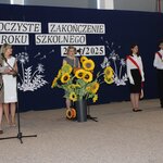 Na uroczystości zakończenia roku szkolnego 2024/2025, kobiety prowadzące mówią do mikrofonu. Obok nich stoi poczet sztandarowy, trzymający sztandar Polski. W tle widoczna dekoracja z napisem „Uroczyste zakończenie roku szkolnego 2024/2025” oraz duża donica ze sztucznymi słonecznikami. Uczniowie, ubrani w białe koszule i czarne spódnice, stoją w postawie honorowej.
